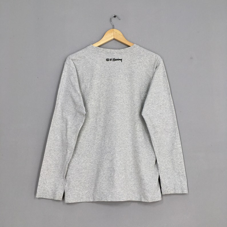 May include: Gray long-sleeve t-shirt with a black print of the artist's signature, "&copy; K. Haring", on the back.