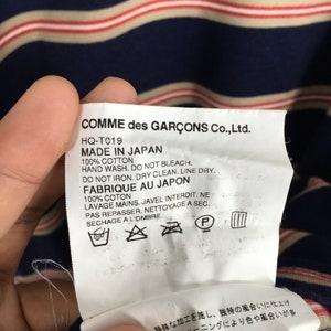 Puede incluir: Una etiqueta de Comme des Gar&ccedil;ons Co., Ltd. con el texto "MADE IN JAPAN" e instrucciones de lavado. La etiqueta se sostiene sobre una tela a rayas azul marino y beige con rayas rojas.
