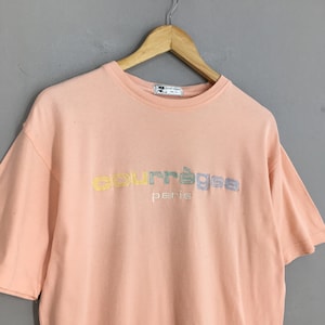 K&ouml;nnte beinhalten: Ein pfirsichfarbenes T-Shirt mit kurzen &Auml;rmeln und dem gestickten Wort "courr&egrave;ges" in Pastellfarben sowie dem Wort "paris" in Wei&szlig;. Das Shirt h&auml;ngt an einem Holzb&uuml;gel.