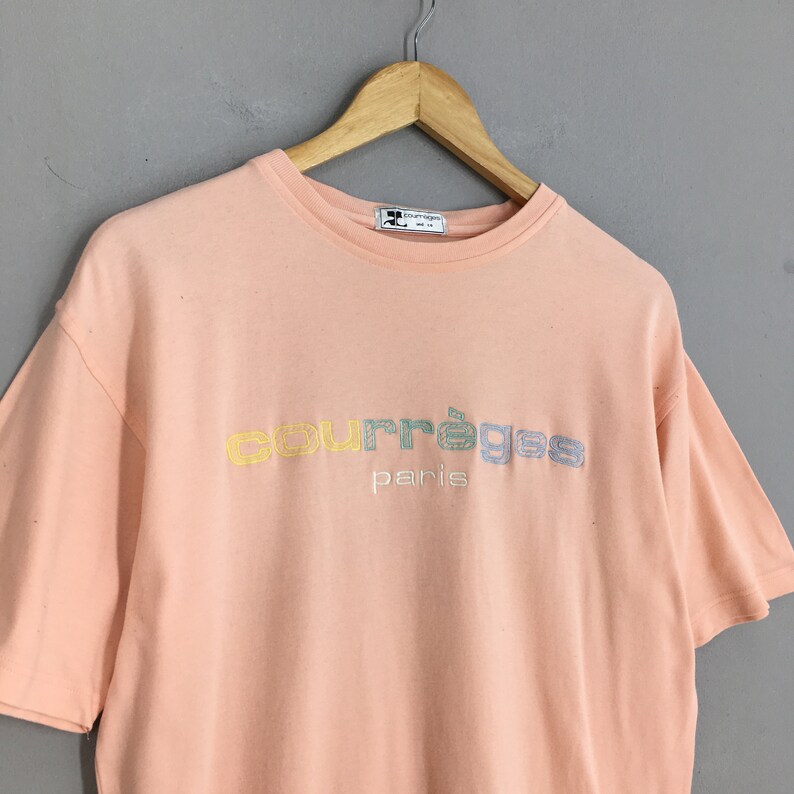 K&ouml;nnte beinhalten: Ein pfirsichfarbenes T-Shirt mit kurzen &Auml;rmeln und dem gestickten Wort "courr&egrave;ges" in Pastellfarben sowie dem Wort "paris" in Wei&szlig;. Das Shirt h&auml;ngt an einem Holzb&uuml;gel.