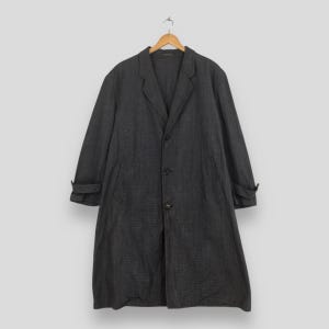 Puede incluir: Un abrigo largo gris carbón con cuello de solapa clásico y tres botones. El abrigo tiene mangas largas con puños doblados y está colgado de una percha de madera. El abrigo está hecho de una tela texturizada.