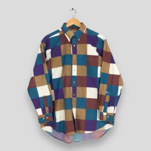 Puede incluir: Una camisa abotonada de manga larga con un diseño de patchwork en tonos turquesa, marrón, morado y blanco. La camisa está colgada de una percha de madera. La camisa tiene un cuello clásico y puños abotonados.