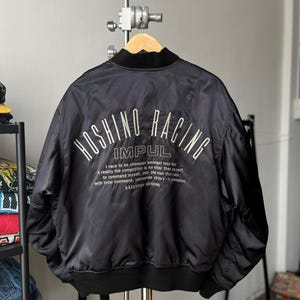 Puede incluir: Chaqueta bomber negra con texto "HOSHINO RACING" bordado en blanco en la espalda. Debajo, el texto "IMPUL" y una cita. La chaqueta tiene cuello y puños de canalé negros. La chaqueta está colgada de una percha de madera.