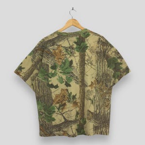 May include: A short-sleeved t-shirt with a camouflage pattern featuring brown tree trunks, green and brown leaves, and branches. The shirt has a green collar and is hanging on a wooden hanger.