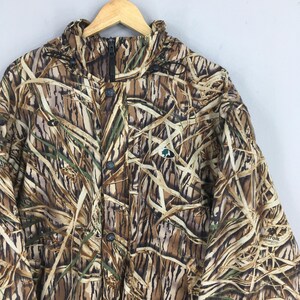 Puede incluir: Chaqueta de camuflaje con un patr&oacute;n marr&oacute;n, beige y verde. La chaqueta tiene cierre de cremallera y botones, cuello alto y un peque&ntilde;o logotipo en el pecho. El patr&oacute;n se asemeja a juncos y hierbas.