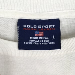 May include: A white Polo Sport Ralph Lauren t-shirt tag with a blue background and white text. The tag says "Polo Sport Ralph Lauren" and "Made in USA 100% Cotton See Reverse for Care". The tag also has a small American flag and a size "L" printed on it.