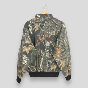Op de afbeelding: Camouflageprintjas met een zwarte rits en een zwarte kraag. De jas heeft een zwarte band rond de onderkant en de woorden "Cabela's" zijn op de achterkant gedrukt.