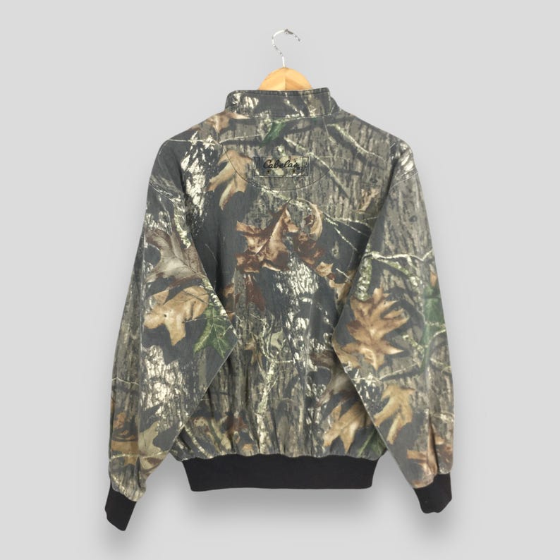 Op de afbeelding: Camouflageprintjas met een zwarte rits en een zwarte kraag. De jas heeft een zwarte band rond de onderkant en de woorden "Cabela's" zijn op de achterkant gedrukt.