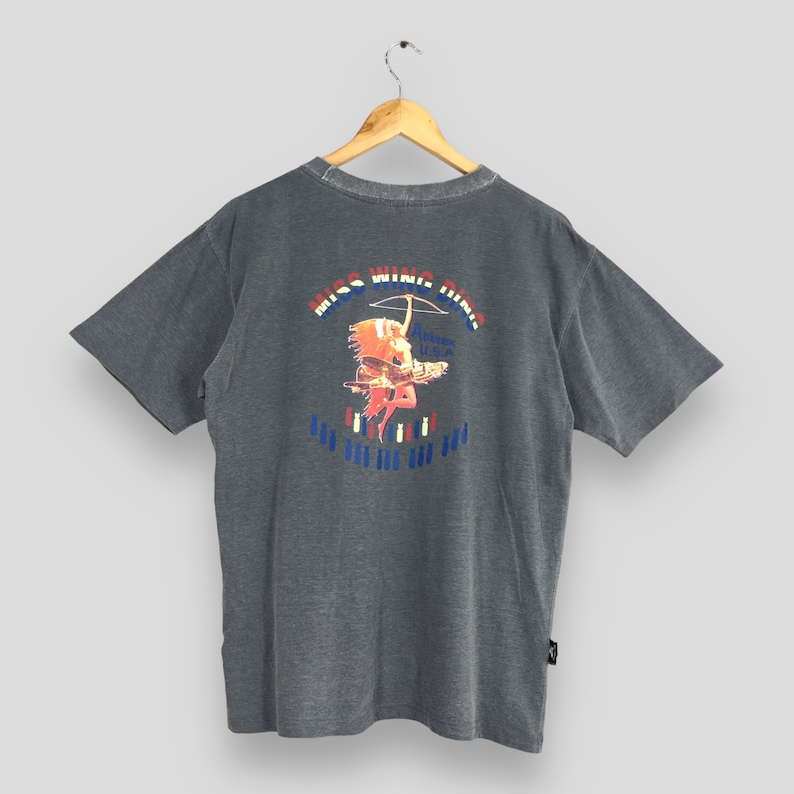 May include: Gray short-sleeve t-shirt with a graphic on the back featuring the text "MISS WING HING" above an illustration of a Native American figure with a bow and arrow. The graphic includes red, white, and blue accents.