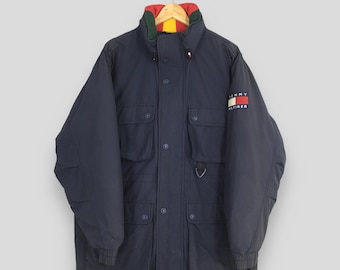 Pode incluir: Casaco de inverno azul marinho com gola multicolorida e o logotipo Tommy Hilfiger na manga. O casaco tem vários bolsos e uma frente abotoada. O casaco está pendurado num cabide de madeira.