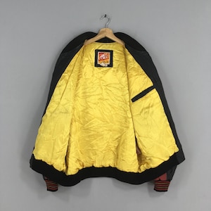 May include: Black bomber jacket with a bright yellow quilted lining. The jacket has a black collar, cuffs, and hem. The cuffs have a red and black striped design. A small patch is visible on the inside of the jacket.