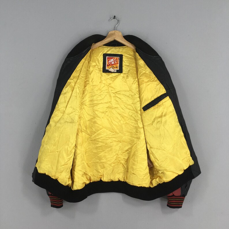 May include: Black bomber jacket with a bright yellow quilted lining. The jacket has a black collar, cuffs, and hem. The cuffs have a red and black striped design. A small patch is visible on the inside of the jacket.