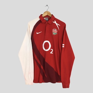 May include: A long-sleeved red and white rugby shirt with a collar, featuring the Nike logo and the England rose emblem. The shirt has the O2 logo and white accents on the sleeves.