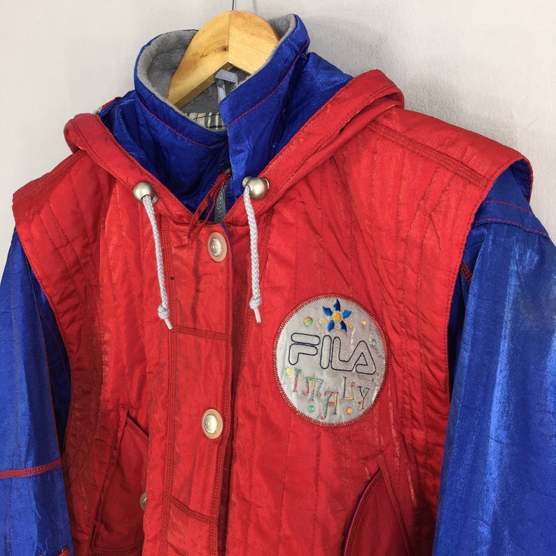 May include: A vintage red and blue Fila jacket with a hood. The jacket features a silver logo patch with the word "ITALY" and the Fila logo. The jacket has a button-up front and a drawstring hood.