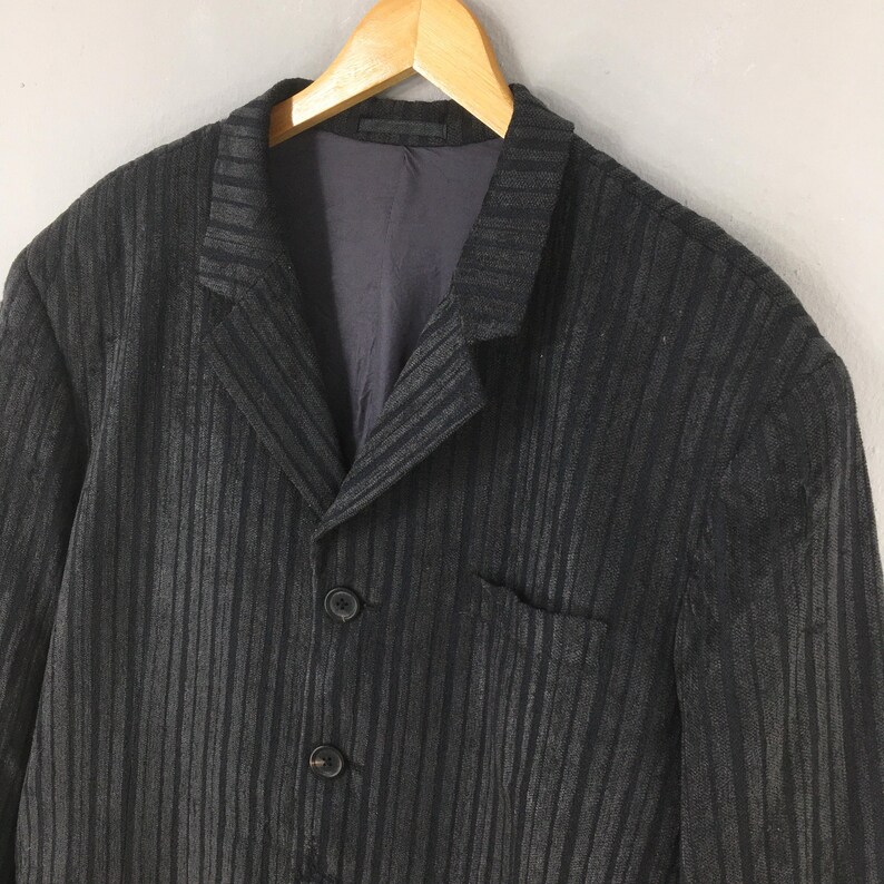 May include: A dark charcoal gray blazer featuring a vertical striped pattern throughout the fabric. The garment includes a notched lapel, a single chest pocket, and two visible buttons on the front closure, all displayed on a wooden hanger.