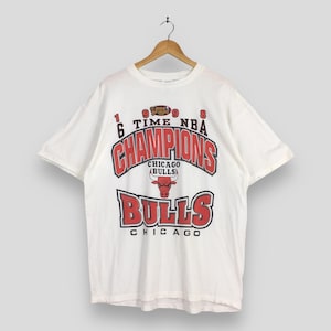 Puede incluir: Camiseta blanca con el logotipo de los Chicago Bulls y las palabras "CHAMPIONS" y "BULLS" en rojo y negro. La camiseta conmemora las victorias de las Finales de la NBA de 1996 y 1998. La camiseta está colgada de una percha de madera.