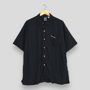 May include: A black short-sleeved button-down shirt with a classic collar and a single chest pocket. The shirt features light-coloured buttons and the word SOLJAH embroidered in white thread on the pocket. It is displayed on a wooden hanger.