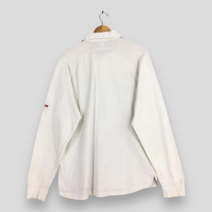 Peut inclure: Chemise blanche &agrave; manches longues avec col, suspendue &agrave; un cintre en bois. La chemise pr&eacute;sente une petite bande rouge sur la manche gauche. La vue arri&egrave;re r&eacute;v&egrave;le le col et les coutures.