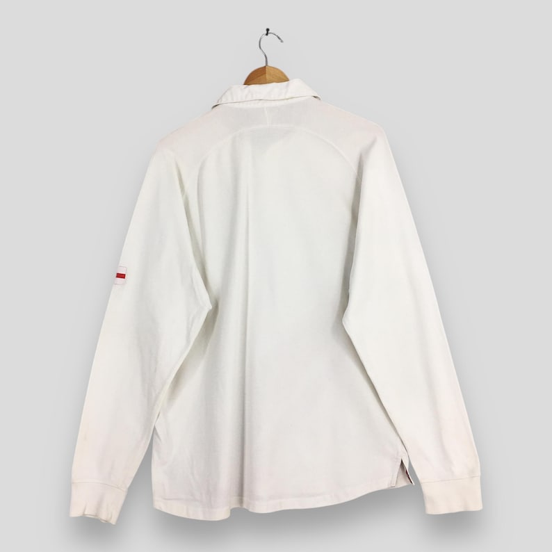 Peut inclure: Chemise blanche &agrave; manches longues avec col, suspendue &agrave; un cintre en bois. La chemise pr&eacute;sente une petite bande rouge sur la manche gauche. La vue arri&egrave;re r&eacute;v&egrave;le le col et les coutures.