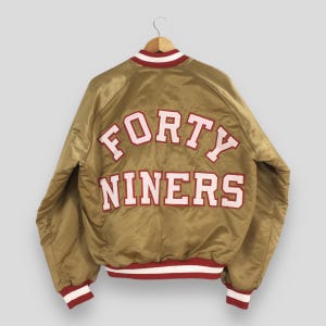 May include: A gold satin varsity jacket featuring the text FORTY NINERS in white letters with red outlines across the back. The jacket has a ribbed collar, cuffs, and hem with red and white stripes, long sleeves, and a button-up front closure.