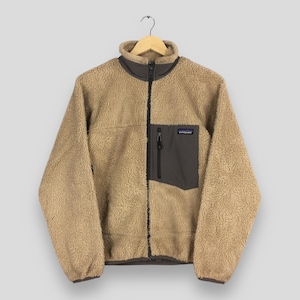 Pode incluir: Um casaco de lã polar bege com gola alta e fecho de correr frontal preto a todo o comprimento. O casaco apresenta um bolso no peito cinzento escuro com fecho vertical e uma etiqueta da marca Patagonia. Os punhos e a bainha têm acabamentos em cinzento escuro.