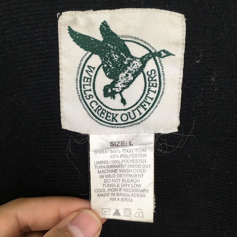 Może przedstawiać: Zbliżenie metki odzieżowej Wells Creek Outfitters. Biała metka z zielonym logo lecącej kaczki. Tekst zawiera rozmiar, szczeg&oacute;ły materiału i instrukcje prania. Czarny materiał w tle.