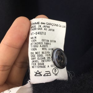 Puede incluir: Etiqueta de cuidado blanca con texto negro sobre fondo negro. La etiqueta dice "COMME des GAR&Ccedil;ONS Co., Ltd. MADE IN JAPAN FABRIQUE AU JAPON HT-040210 MAIN 100% COTTON COTON ATTACHED FABRIC TISSU AJOUTE 100% WOOL LAINE DO NOT BLEACH USE COOL IRON DRY CLEAN ONLY JAVEL INTERDIT FER DOUX NETTOYAGE A SEC" con un bot&oacute;n negro y s&iacute;mbolos de cuidado.