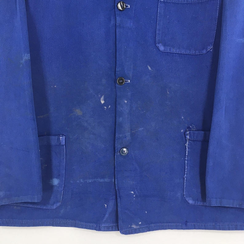 Peut inclure: Gros plan sur une veste de travail bleue avec trois boutons visibles et une poche. La veste pr&eacute;sente des &eacute;claboussures de peinture et des marques d'usure. Fabriqu&eacute;e dans un mat&eacute;riau r&eacute;sistant.
