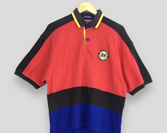 May include: A color-blocked polo shirt in red, black, and blue. The shirt has a yellow and gray collar with a small logo patch on the chest. The short sleeves are black.