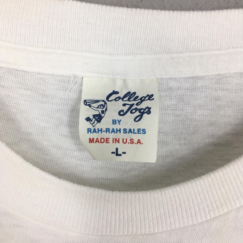 May include: Close-up of a white t-shirt collar with a rectangular tag. The tag features the text "College Jogs" in a blue script font, along with "BY RAH-RAH SALES MADE IN U.S.A." and the size "-L-".