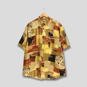 以下が含まれることがあります： ベージュ、茶色、黄色、黒の色合いの幾何学模様の半袖シャツ。 シャツは襟付きで、胸ポケットがあり、木製のハンガーに吊るされています。