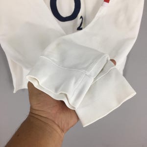 Peut inclure: T-shirt blanc &agrave; manches longues avec lettrage bleu marine et le chiffre 2. Le t-shirt a un poignet c&ocirc;tel&eacute; et un d&eacute;tail de rayure rouge. Le tissu semble &ecirc;tre un mat&eacute;riau tricot&eacute; doux.