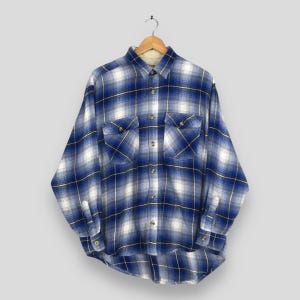 Puede incluir: Camisa de franela a cuadros azules y blancos con dos bolsillos en el pecho y puños abotonados. La camisa de manga larga está colgada de una percha de madera. Tiene un estilo clásico e informal.