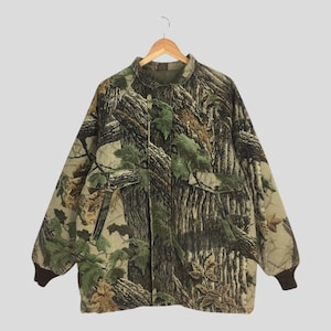 May include: A camouflage jacket featuring a tree bark and leaf design. The jacket has a button-up front, two front pockets, and brown ribbed cuffs. The jacket is suspended on a wooden hanger.
