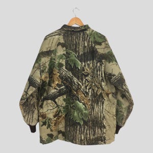 May include: A camouflage jacket with a button-down collar and brown cuffs. The jacket features a pattern of tree bark and green leaves, designed to blend into a forest environment. The jacket is hanging on a wooden hanger.