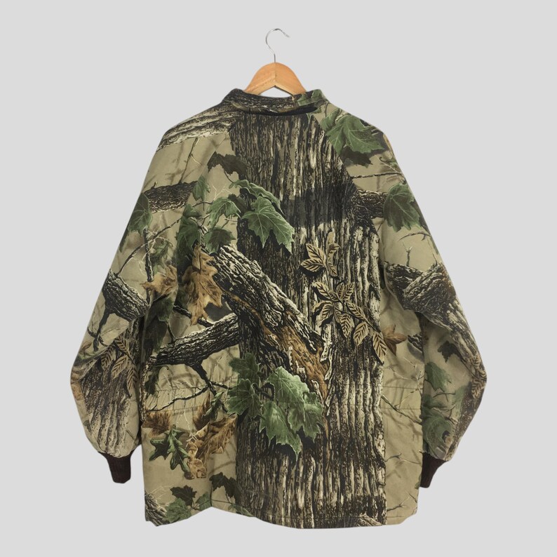 May include: A camouflage jacket with a button-down collar and brown cuffs. The jacket features a pattern of tree bark and green leaves, designed to blend into a forest environment. The jacket is hanging on a wooden hanger.