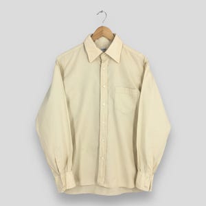 Pode incluir: Uma camisa de manga comprida de cor bege claro com um bot&atilde;o e um bolso no peito. A camisa tem uma gola cl&aacute;ssica e um fecho de bot&otilde;es na frente.