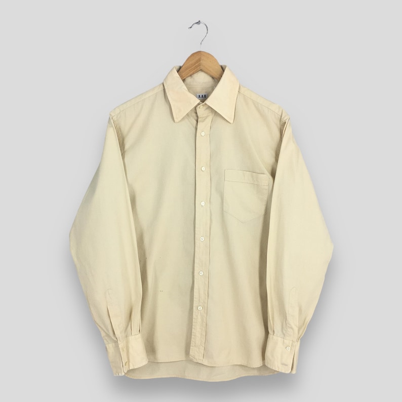 Pode incluir: Uma camisa de manga comprida de cor bege claro com um bot&atilde;o e um bolso no peito. A camisa tem uma gola cl&aacute;ssica e um fecho de bot&otilde;es na frente.