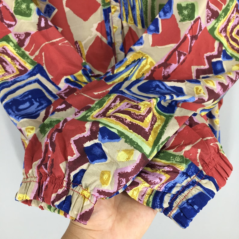 May include: A red, blue, yellow, green, and brown patterned shirt with a geometric design. The shirt has a button-up front and long sleeves with elastic cuffs.