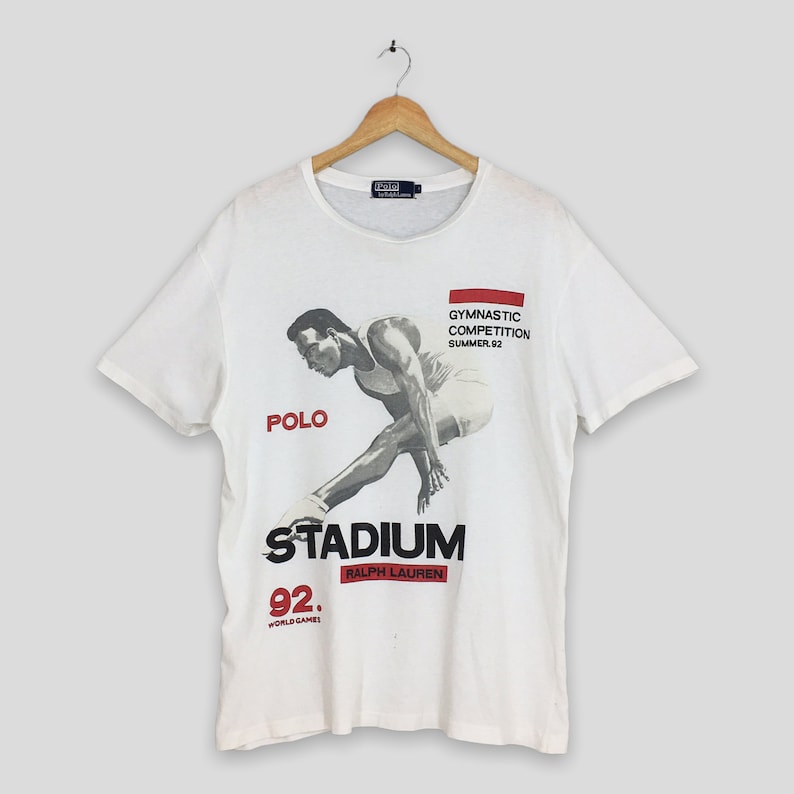 May include: White t-shirt featuring a graphic print of a man performing a gymnastic pose. The graphic includes the text "Polo", "Stadium", "Ralph Lauren", "Gymnastic Competition Summer 92", and "92 World Games".