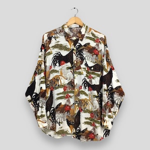 Puede incluir: Camisa de manga larga con estampado de gallos. La camisa presenta un fondo blanco con un patrón repetido de gallos en varios colores, incluyendo negro, marrón y rojo. Cuelga de una percha de madera.