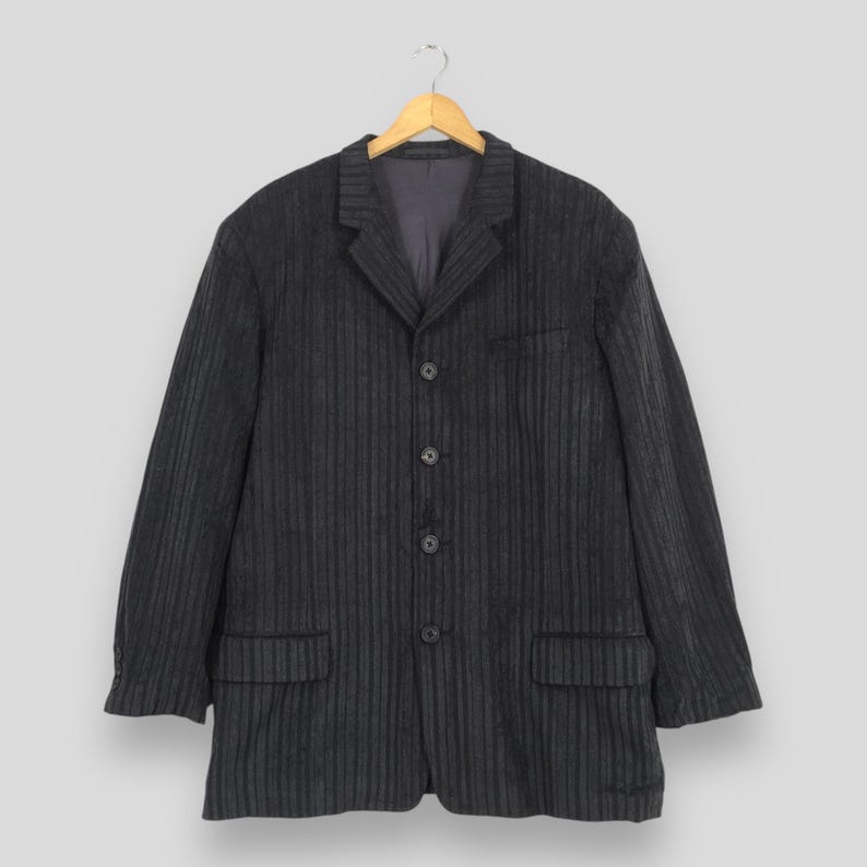 May include: A dark charcoal gray blazer featuring a subtle vertical pinstripe pattern. This garment includes a notched lapel, a four-button front closure, two lower flap pockets, and long sleeves with buttoned cuffs, all displayed on a wooden hanger.