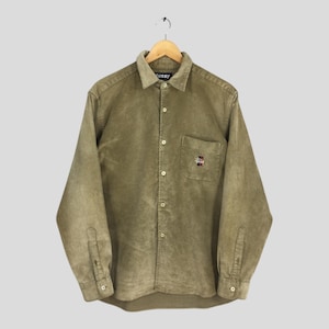 May include: A long-sleeved, button-down corduroy shirt in a muted olive green. The shirt features a small pocket on the chest with a logo and the word "Stussy" on the collar. The shirt is hanging on a wooden hanger.