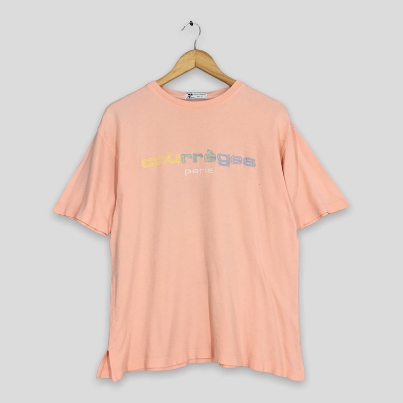 K&ouml;nnte beinhalten: Ein pfirsichfarbenes T-Shirt mit dem Text "courr&egrave;ges paris" in Pastellfarben. Das kurz&auml;rmelige Shirt h&auml;ngt an einem Holzb&uuml;gel. Das Shirt hat eine lockere Passform und einen Schlitz an den Seiten.