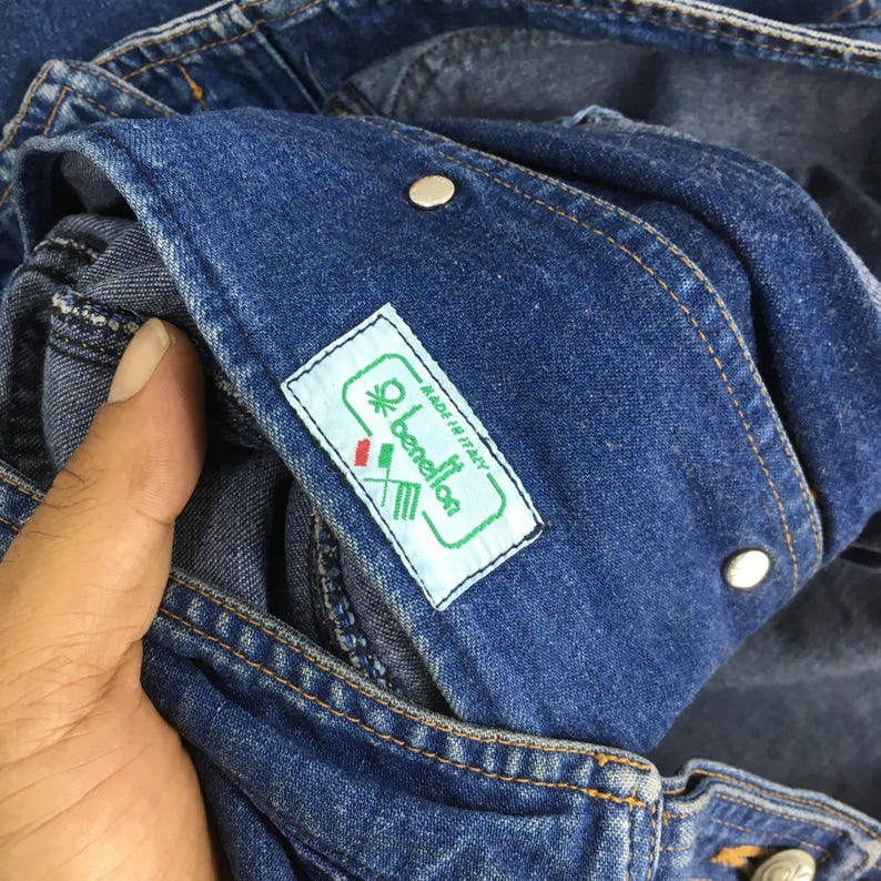 May include: Close-up of blue denim fabric with a label that reads "benetton" and "Made in Italy". The label is white with green and red accents. Two silver buttons are visible.