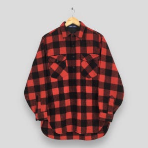 May include: A red and black checked flannel shirt with two chest pockets and buttoned cuffs. The long-sleeved shirt is hanging on a wooden hanger against a neutral background. The shirt has a classic lumberjack style.