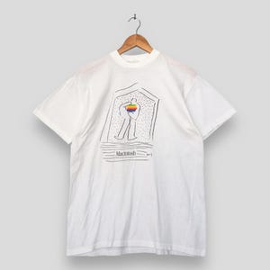 以下が含まれることがあります： 白い半袖Tシャツ。ドアの中に立っている人物の黒い線画が描かれています。人物は虹色の四角い物体を持っています。人物の下にはMacintoshという文字と水平線が黒でプリントされています。