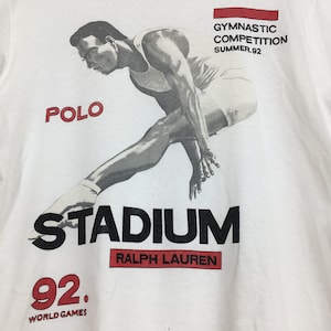 May include: White t-shirt featuring a black and white image of a gymnast in mid-air. The text "STADIUM RALPH LAUREN" is printed in black below the image. The text "92. WORLD GAMES" is printed in red below the text "STADIUM". The text "POLO" is printed in red in the top left corner of the image. The text "GYMNASTIC COMPETITION SUMMER '92" is printed in black in the top right corner of the image.