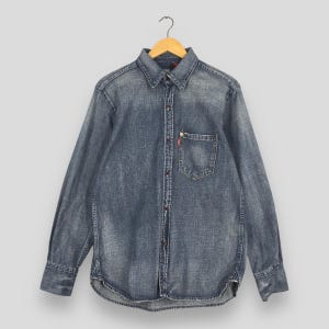 May include: A long-sleeve denim shirt in a faded blue wash featuring a standard collar, a button-down front, and a single chest pocket with a small red tab. The shirt is displayed on a wooden hanger and has a curved hemline at the bottom.