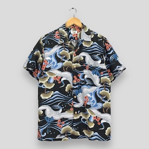 Puede incluir: Camisa negra de manga corta con un estampado de grullas y olas. La camisa presenta grullas blancas, olas azules y detalles en naranja y verde. La camisa está colgada en una percha de madera.